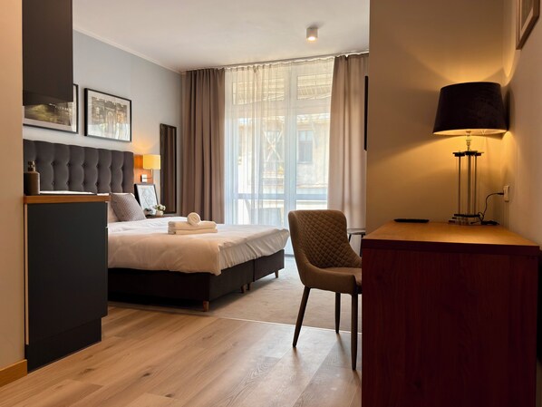 Une chambre d’hôtel avec un lit, un bureau, une chaise, une lampe et une fenêtre avec des rideaux.