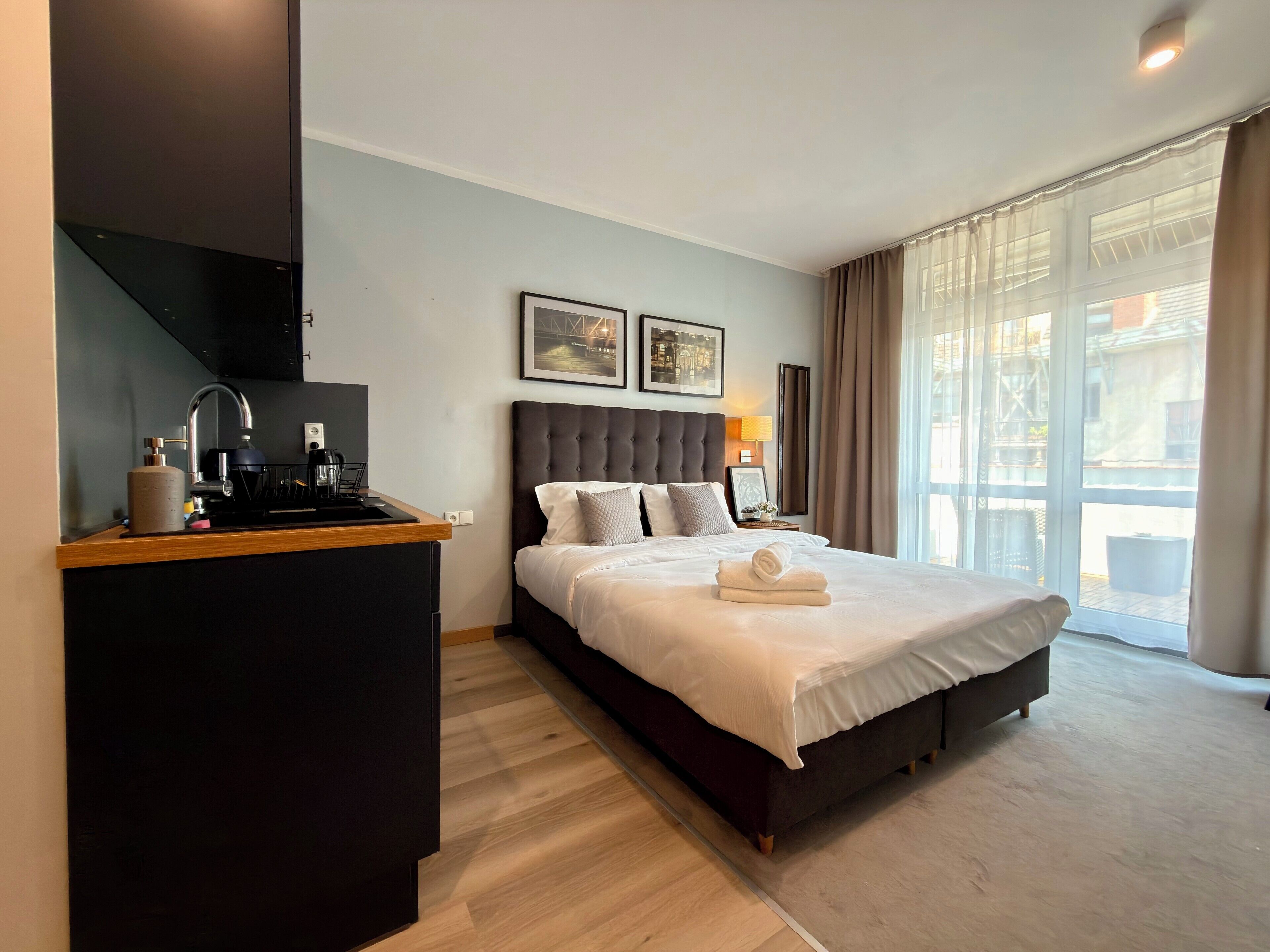 A hotel room with a bed, a small kitchenette, a window with curtains, and two framed pictures on the wall.