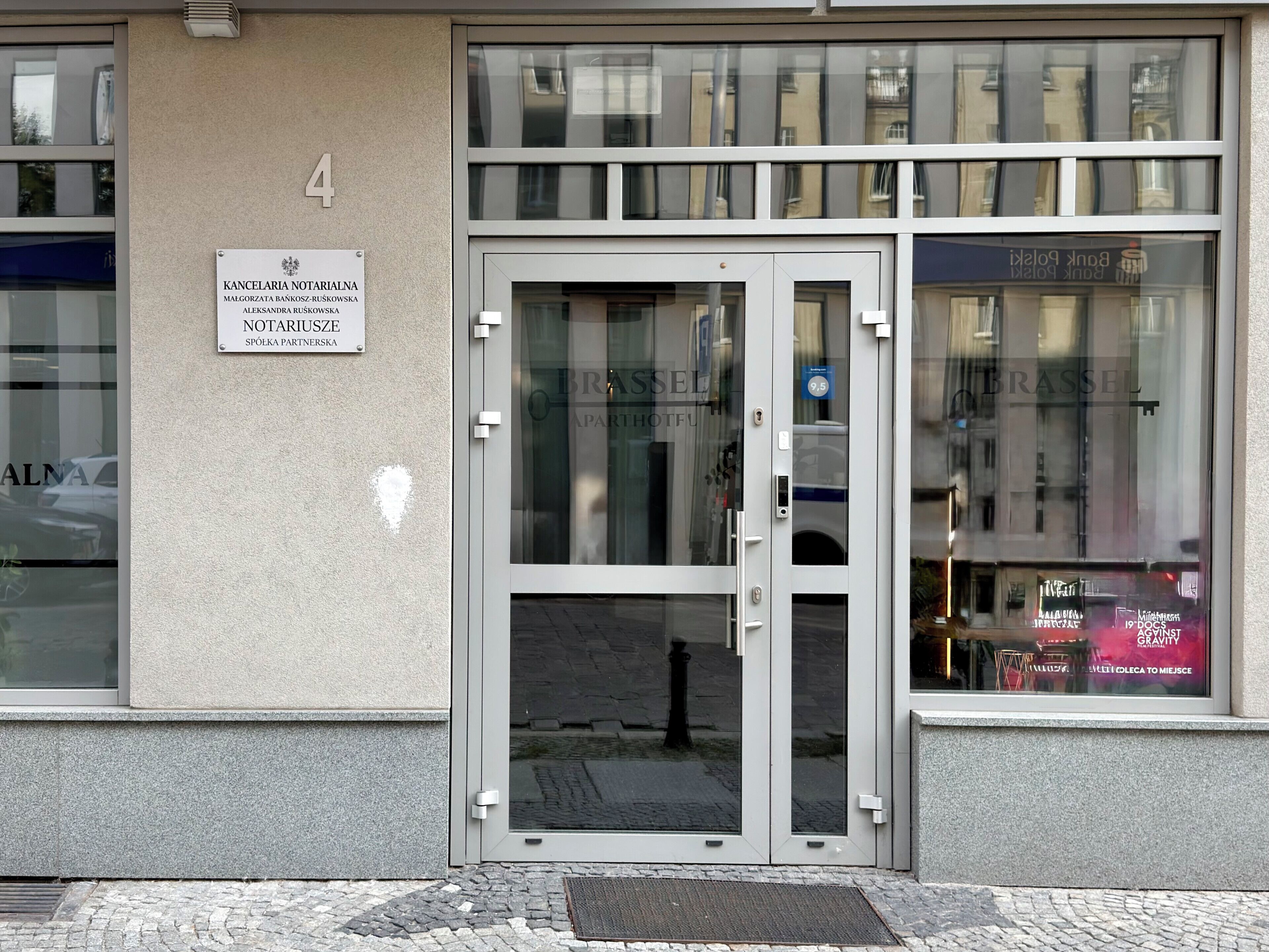Une entrée d’immeuble avec une porte en verre, une enseigne au-dessus de la porte et le reflet d’un autre immeuble visible à travers le verre.