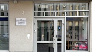 Une entrée d’immeuble avec une porte en verre, une enseigne au-dessus de la porte et le reflet d’un autre immeuble visible à travers le verre.