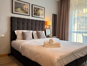 Une chambre d’hôtel avec un grand lit, une tête de lit, deux tableaux encadrés au mur, une lampe de chevet et une fenêtre avec des rideaux.