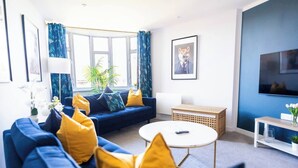 A modern living room with a blue sofa, yellow cushions, a round coffee table, a television, and a large window with curtains. - #141 Coastal Apartment BournemouthBeach| Sleeps 10 (Bournemouth)