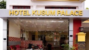 Entrada de hotel com uma placa indicando 'HOTEL KUSUM PALACE', área de recepção com um sofá vermelho, uma mesa de centro e plantas em vasos.