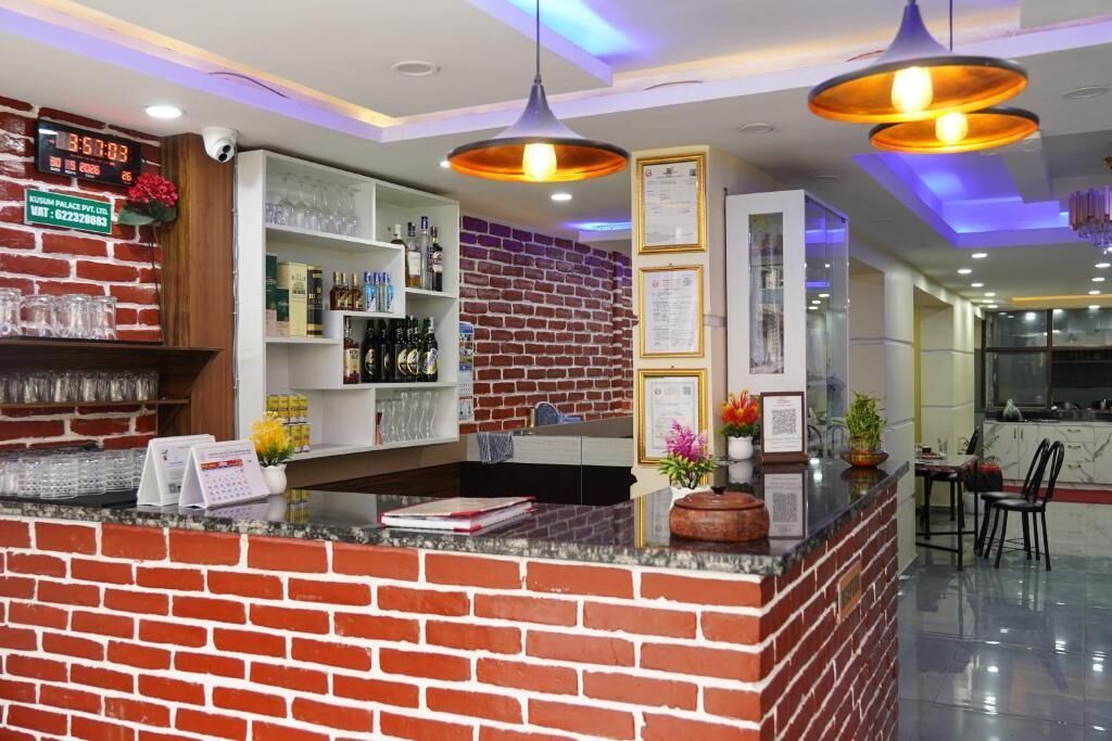 Zona de recepción de un hotel con mostrador de ladrillo, estantes con diversos objetos, un reloj digital e iluminación colgante.