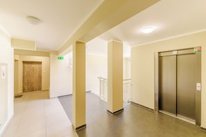 Un couloir avec un ascenseur, une porte en bois et un panneau indiquant la sortie de secours.