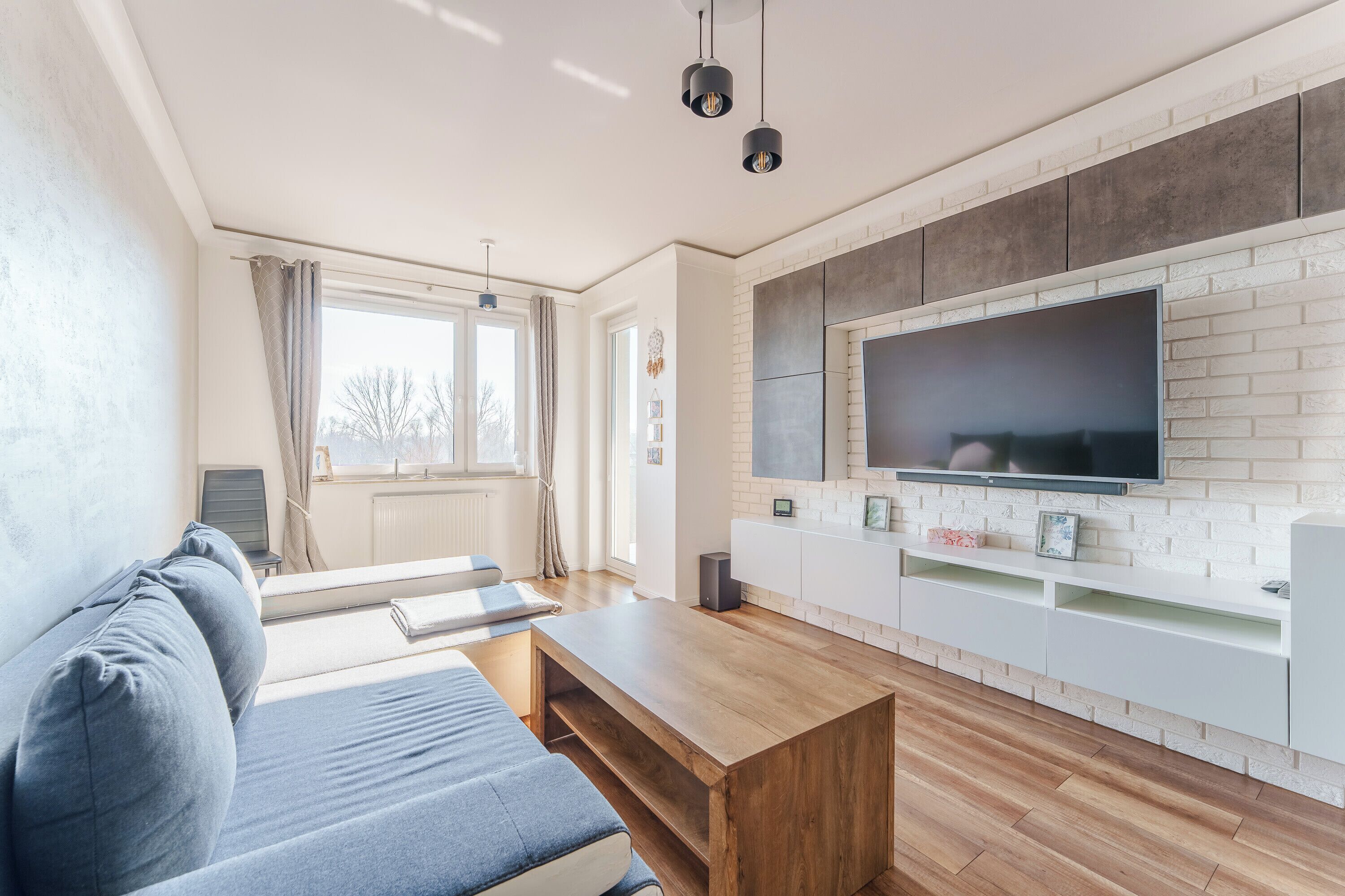 Un salon moderne comprenant un canapé, une table basse, un téléviseur fixé au mur, qui est décoré d’un motif en briques, et une grande fenêtre avec des rideaux.