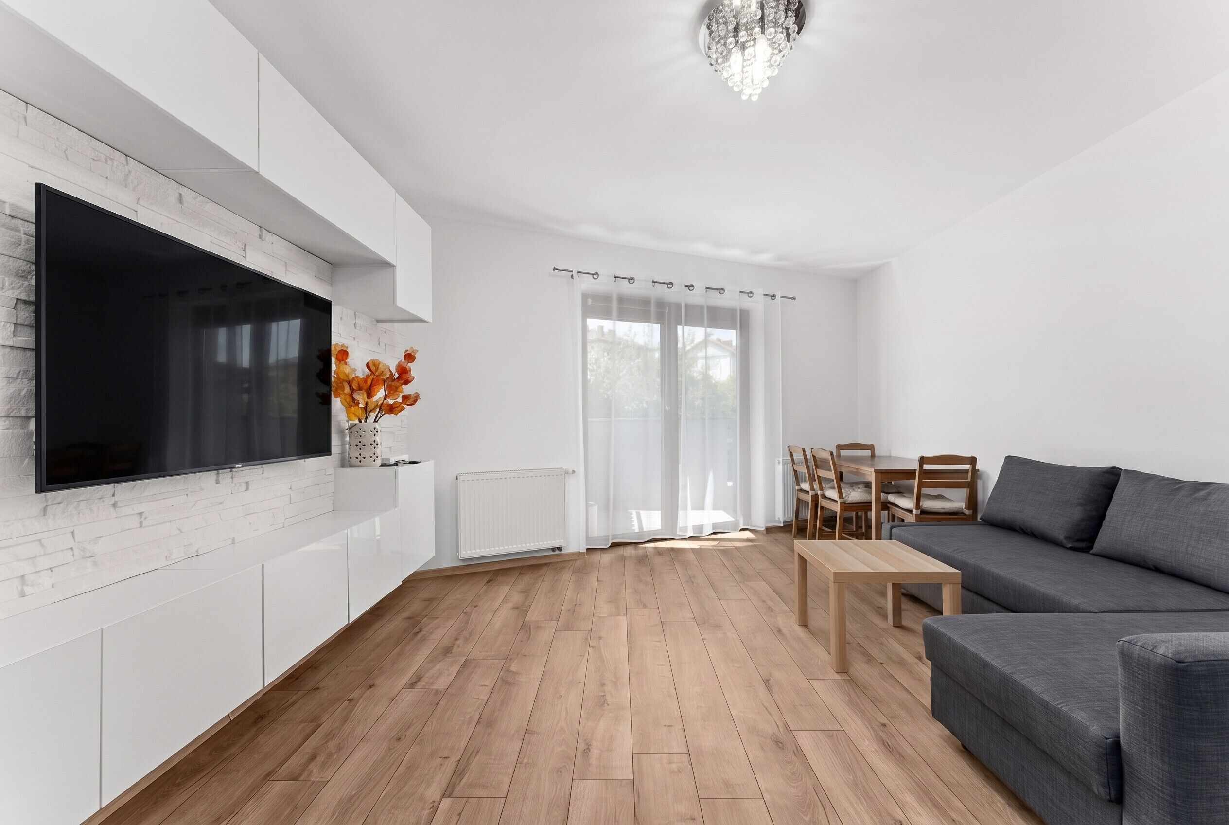 Un soggiorno moderno con TV a schermo piatto montata su una parete di mattoni bianchi, un divano grigio, un tavolo da pranzo in legno con sedie e un'ampia finestra con tende leggere.