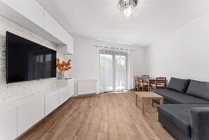 Una sala moderna con un televisor de pantalla plana montado en una pared de ladrillo blanco, un sofá gris, una mesa de comedor de madera con sillas y un amplio ventanal con cortinas traslúcidas.