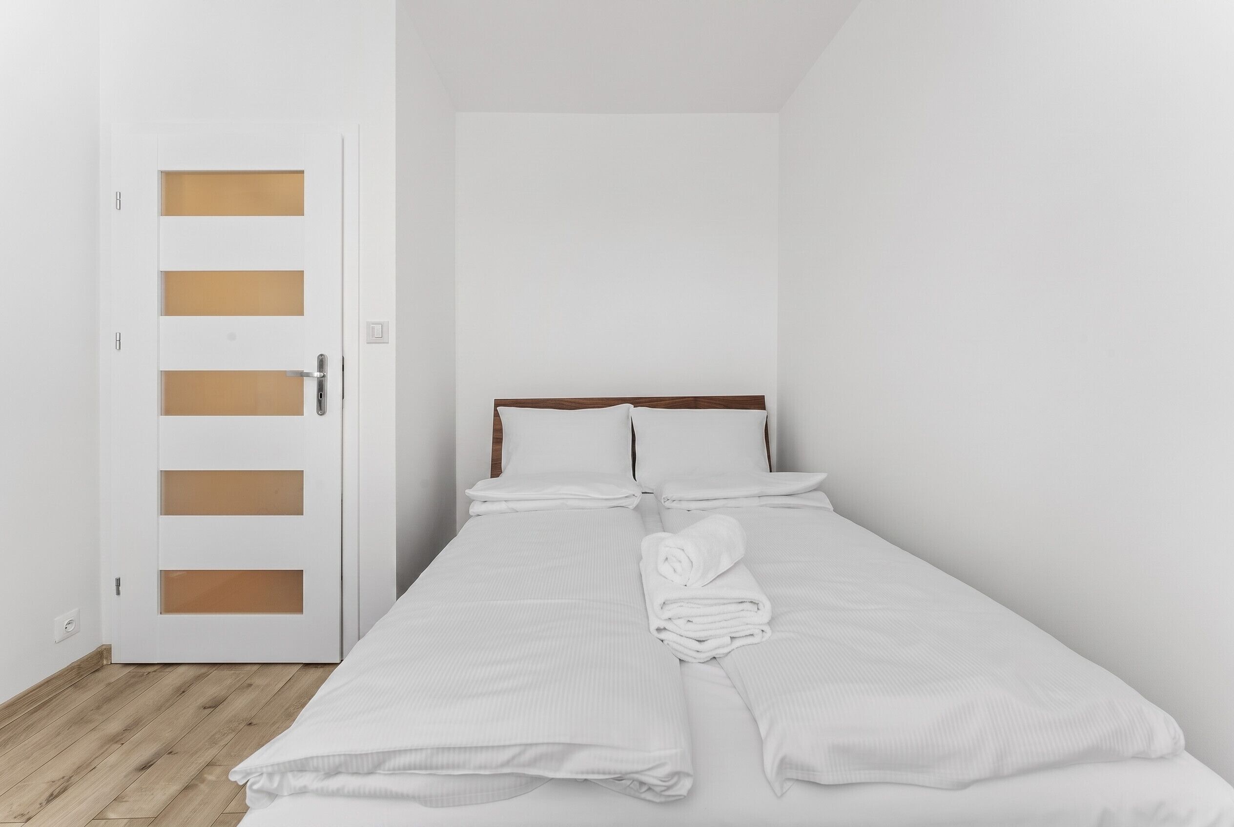 Un letto singolo con biancheria bianca e un asciugamano arrotolato, una porta di legno con maniglia e un interruttore sulla parete.