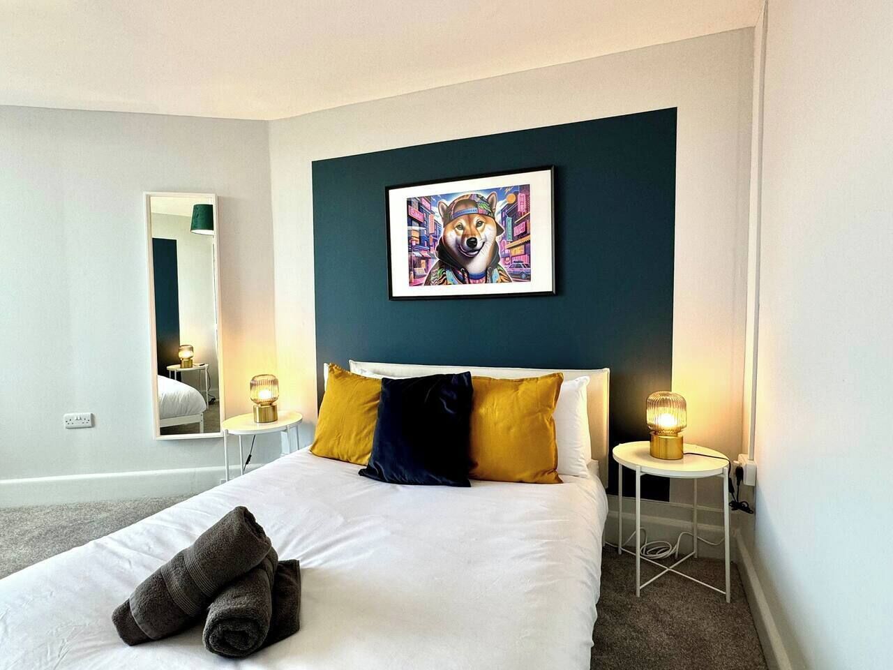 A hotel room with a bed, a bedside table with a lamp, a wall-mounted mirror, and a framed picture above the bed.