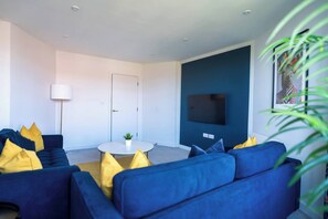 Ein modernes Wohnzimmer mit einem blauen Sofa, einem runden Couchtisch und einem an der Wand befestigten Flachbildfernseher.