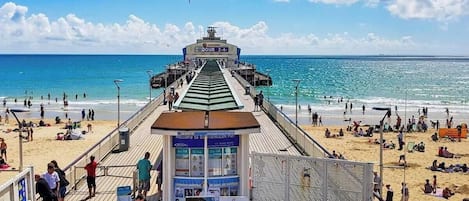 A pier with a ticket booth and information signs, leading to a beach with people and umbrellas.