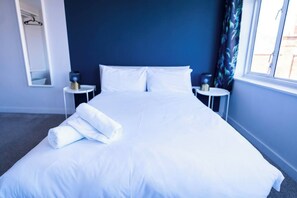A neatly made bed with white linens and a rolled towel, a bedside table with a lamp, and a window with curtains. - #142 Bournemouth Flat| Near AlumChine Beach|for 10 (Bournemouth)