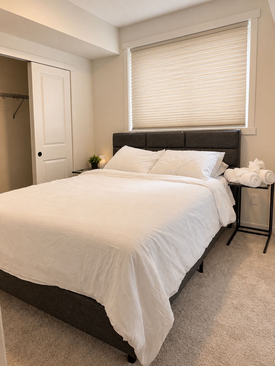 A bedroom with a bed, a bedside table with a lamp, a window with blinds, and a door.