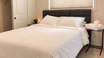 A bedroom with a bed, a bedside table with a lamp, a window with blinds, and a door.