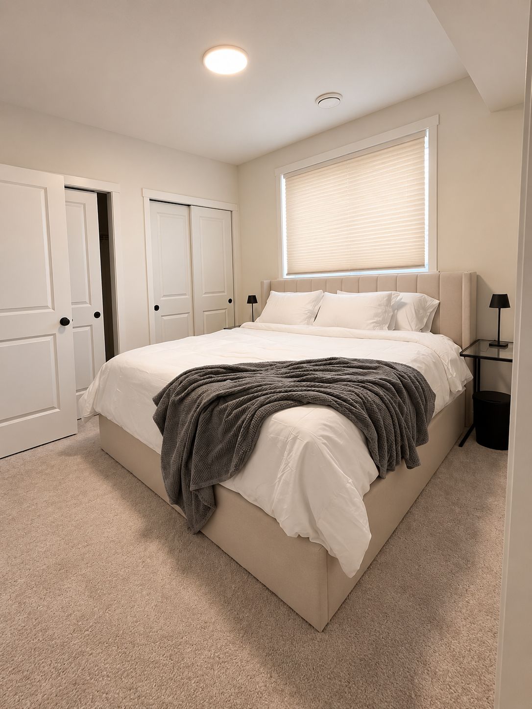 A bedroom with a bed, a window with blinds, a door, and a nightstand with a lamp.
