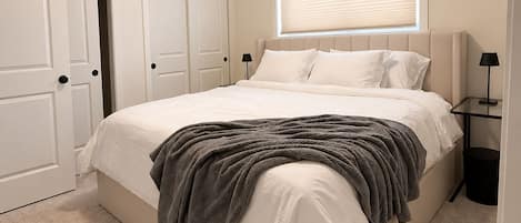 A bedroom with a bed, a window with blinds, a door, and a nightstand with a lamp.