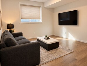 A modern living room with a sofa, a coffee table, a TV mounted on the wall, and a window with blinds. - Modern 2BR Retreat | King Bed | Heated Bath Floors (Edmonton)