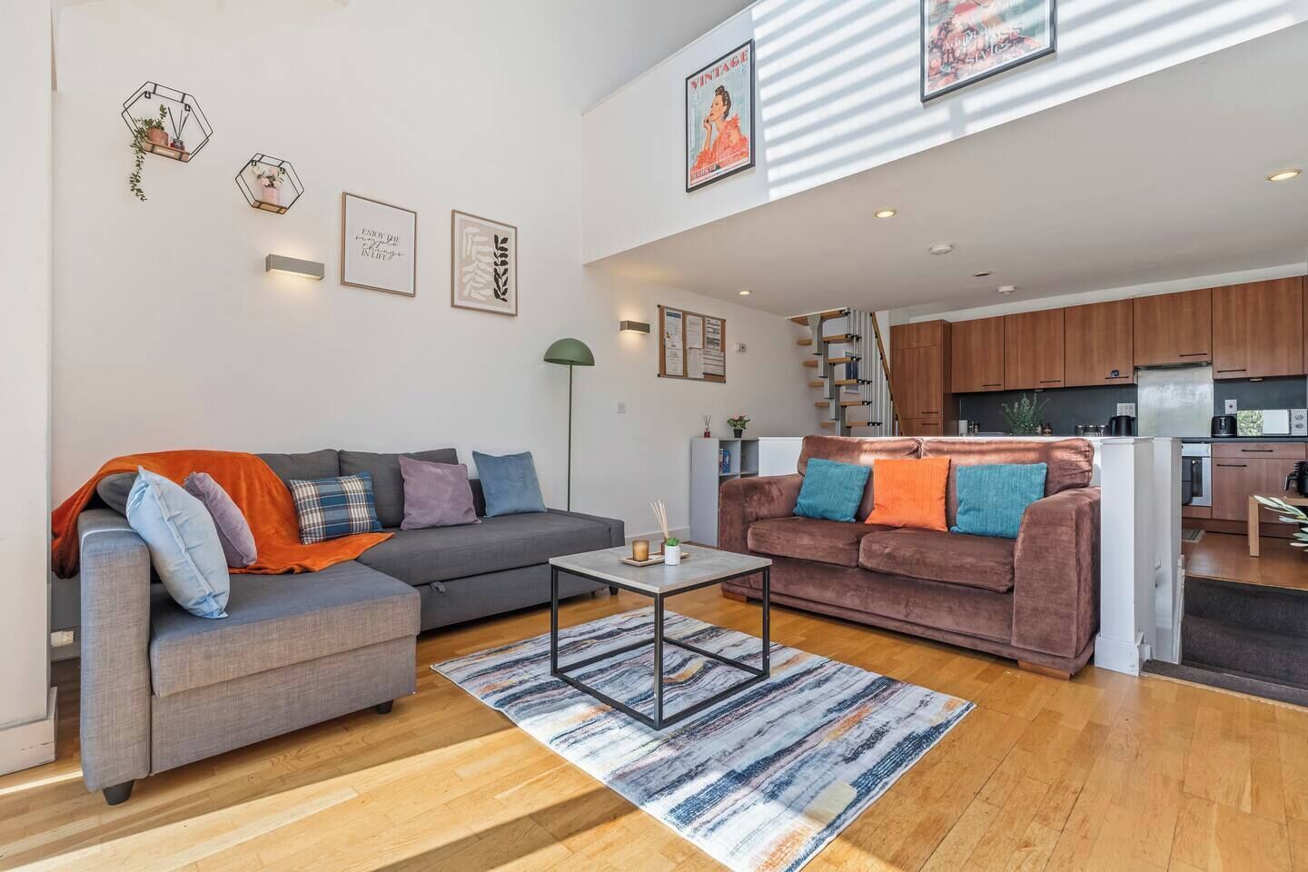 A modern living room with a grey sofa, a brown sofa, and a coffee table. There is a kitchen in the background with wooden cabinets and a white refrigerator.
