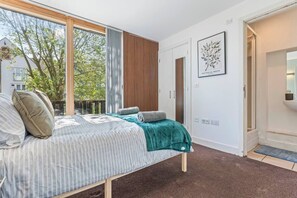 Een slaapkamer met een groot bed, uitzicht op bomen, een deur naar de badkamer en een ingelijst botanisch kunstwerk aan de wand.