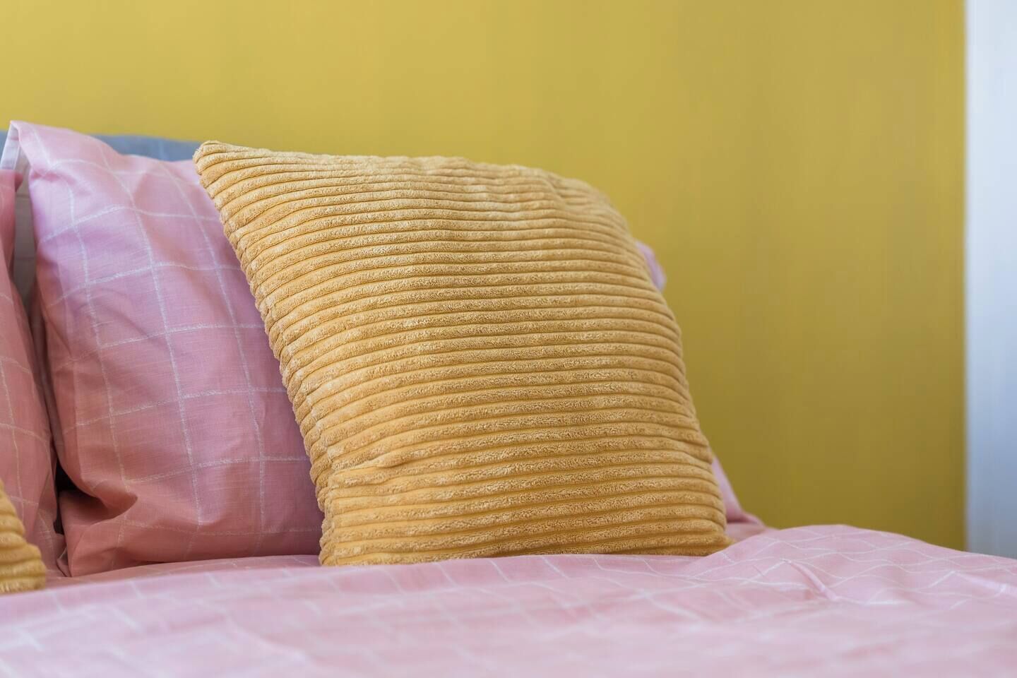 Cama com travesseiro amarelo texturizado e um travesseiro xadrez rosa, tudo contra uma parede amarela.