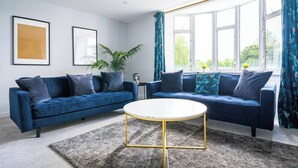 A modern living room with two blue sofas, a round coffee table, and a large window with curtains. - #158 Cozy Flat Near Bournemouth Beach - Sleeps 10 (Bournemouth)