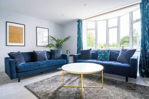 A modern living room with two blue sofas, a round coffee table, and a large window with curtains. - #158 Cozy Flat Near Bournemouth Beach - Sleeps 10 (Bournemouth)