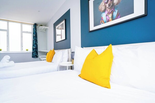 Ein Hotelzimmer mit zwei Betten, weißer Bettwäsche, gelben Kissen, einem gerahmten Bild an der Wand und einem Fenster mit Vorhängen.