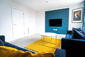 Una sala de estar moderna con un sofá, una mesa de centro, un televisor de pantalla plana montado en una pared azul y una puerta con pomo dorado.