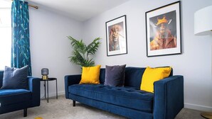 Ein Wohnzimmer mit einem blauen Sofa, gelben Kissen, einer Topfpflanze und zwei gerahmten Bildern an der Wand.