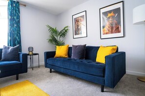 Ein Wohnzimmer mit einem blauen Sofa, gelben Kissen, einer Topfpflanze und zwei gerahmten Bildern an der Wand.