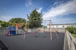 Ein Spielplatz mit Schaukeln und Rutsche, umzäunt und von Bäumen gesäumt.