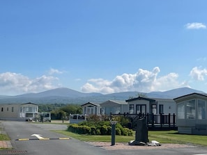 A caravan park with multiple caravans, a paved road, and a mountain in the background. - #155 Cozy Caravan in Dinas Dinlle - Sleeps 6 (Dinas Dinlle)