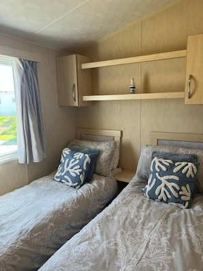 A bedroom with two beds, each with patterned bedding and pillows, a window with curtains, and a wooden shelf above the beds.