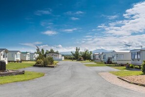 A row of mobile homes with a paved road in the center, surrounded by greenery and a clear sky. - #155 Cozy Caravan in Dinas Dinlle - Sleeps 6 (Dinas Dinlle)