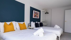 A hotel room with two beds, white bedding, a blue accent wall, a painting, a bedside table with a lamp, and a wardrobe. - #150 Bright Apt – Westbourne Area - Sleeps 7 (Bournemouth)