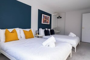 A hotel room with two beds, white bedding, a blue accent wall, a painting, a bedside table with a lamp, and a wardrobe. - #150 Bright Apt – Westbourne Area - Sleeps 7 (Bournemouth)
