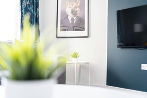 Una habitación con un escritorio blanco, una pequeña mesa blanca con una planta, un cuadro en la pared y un televisor fijado a la pared.