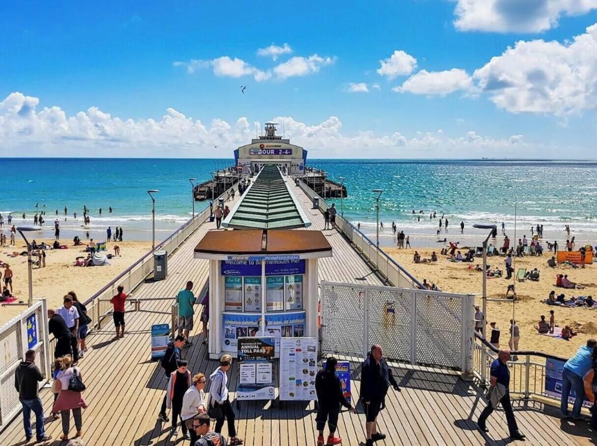 Een steiger met een kaartverkoopkantoor en informatieborden, die uitkomt op een strand met mensen en parasols.