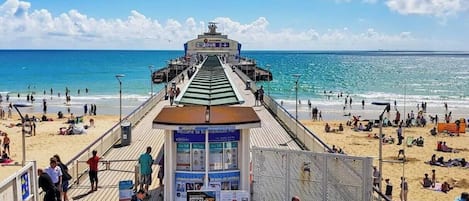 A pier with a ticket booth and information signs, leading to a beach with people and umbrellas.