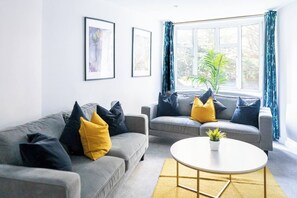 Ein modernes Wohnzimmer mit zwei grauen Sofas, gelb-schwarzen Kissen, einem runden Couchtisch und einem großen Fenster mit Vorhängen.