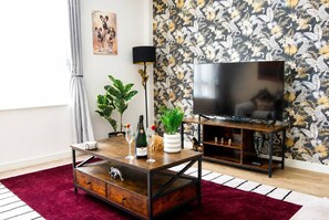 A living room with a television, a coffee table with a bottle and glasses, a plant, and a red rug. - #146 Urban Stay at Sapphire Court - Sleeps 6 (West Midlands)