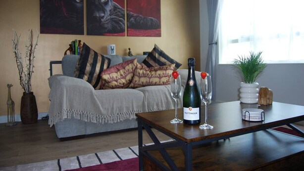 A living room with a sofa, a table with a bottle and glasses, and a window with curtains. - #146 Urban Stay at Sapphire Court - Sleeps 6 (West Midlands)