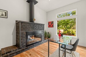 Ein modernes Wohnzimmer mit Kamin aus Stein, einem Glastisch und Blick auf einen Garten durch ein großes Fenster.