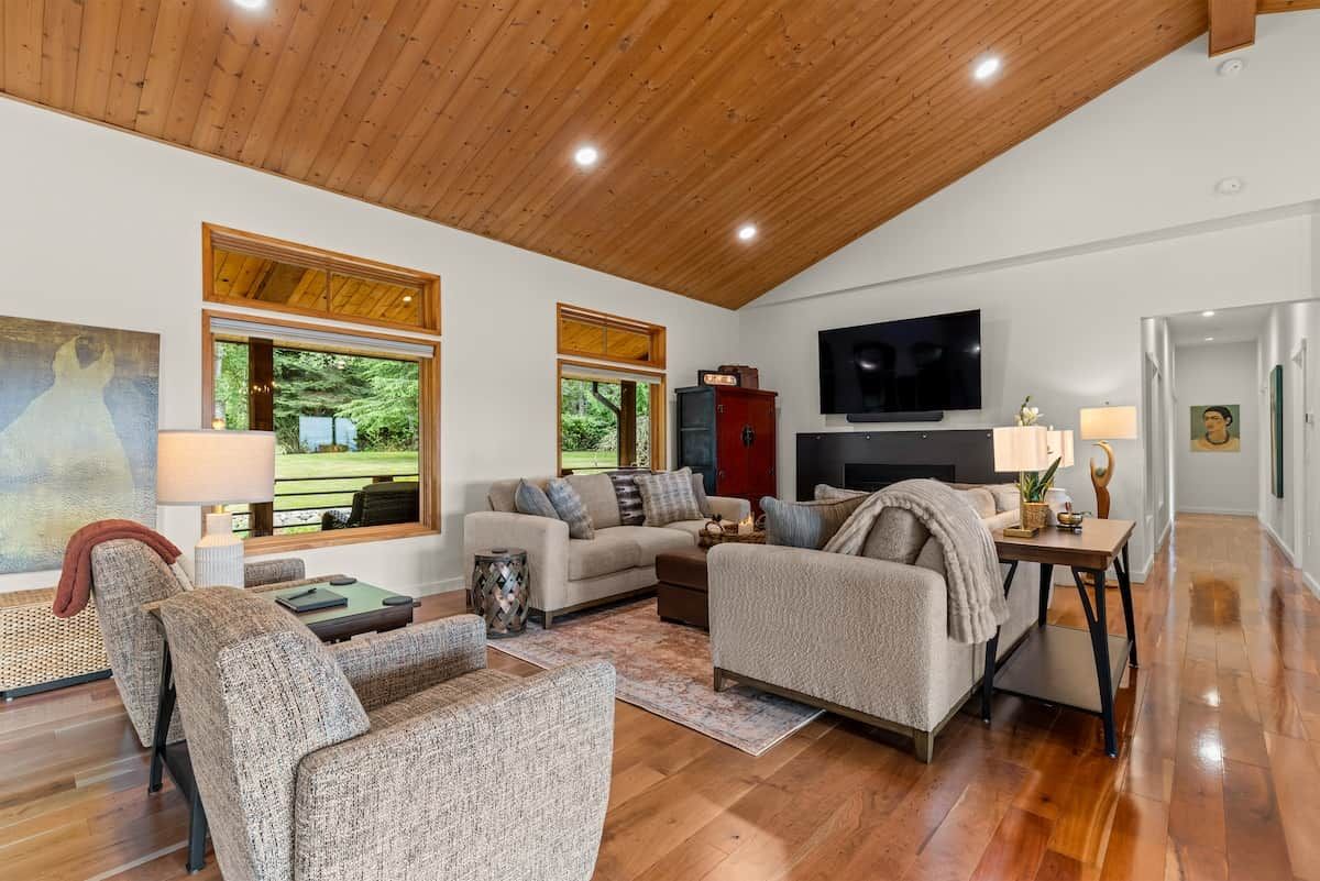 Een ruime woonkamer met een hoge plafond, houten vloeren en grote ramen met uitzicht op groen.