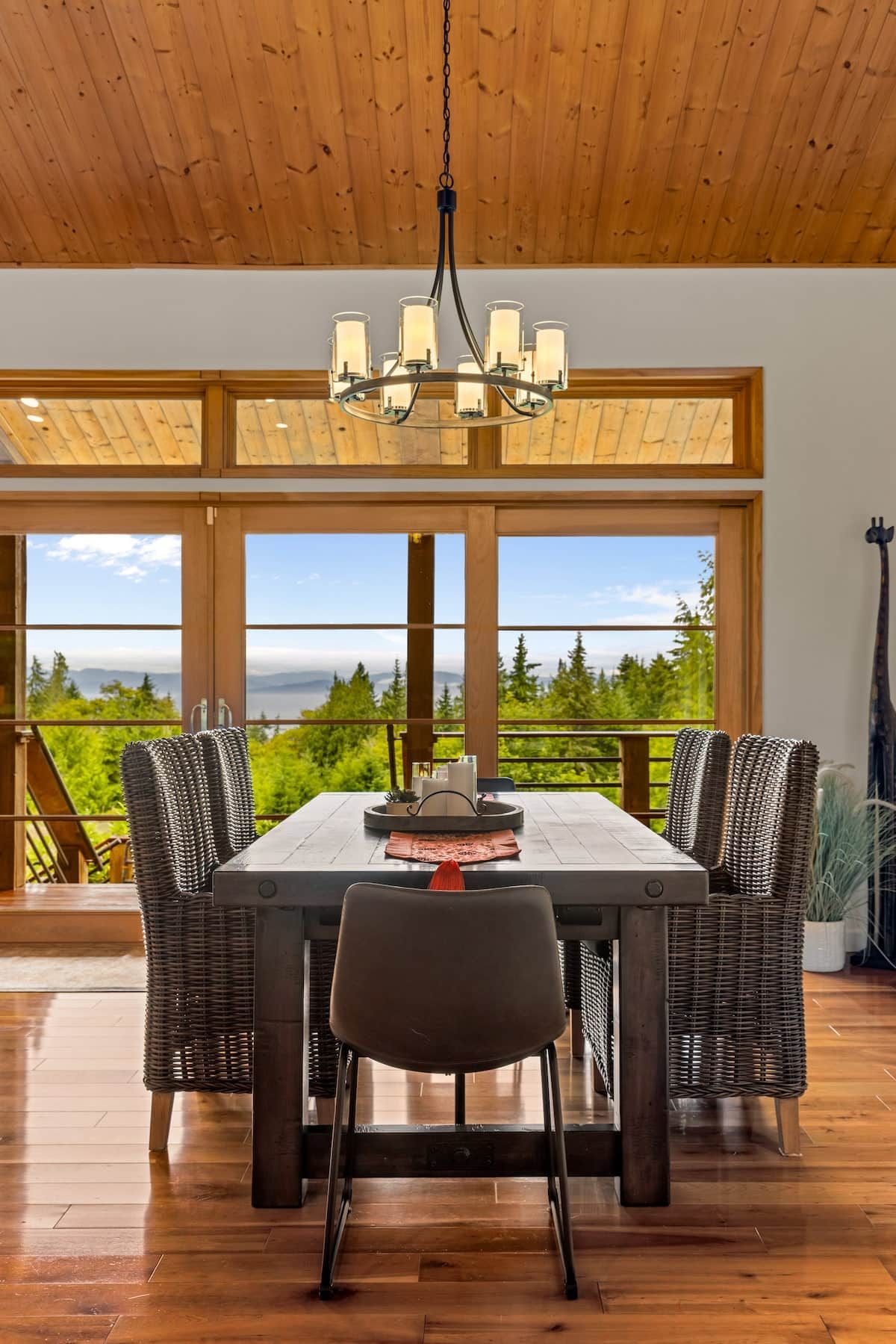 Un coin repas avec une table en bois, des chaises et un lustre. De grandes fenêtres offrent une vue sur la verdure et les montagnes.