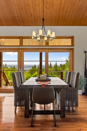 A dining area with a wooden table, chairs, and a chandelier. Large windows offer a view of greenery and mountains. - The Residence at Freshwater Bay (Port Angeles)