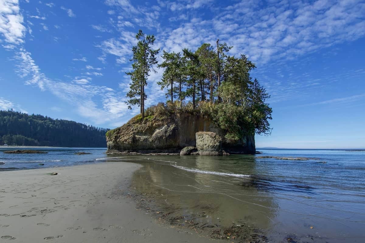 A natural landscape with a sandy beach, calm water, and a rocky outcrop with trees.