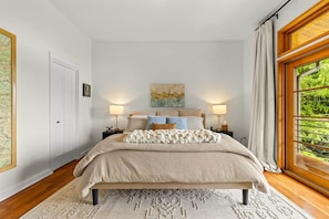 A bedroom with a large bed, two bedside tables, a nightstand, a window with curtains, and a wall-mounted painting. - The Residence at Freshwater Bay (Port Angeles)