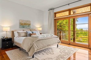 Quarto com cama, abajures, tapete e uma grande porta de vidro deslizante que dá para uma varanda com vegetação.